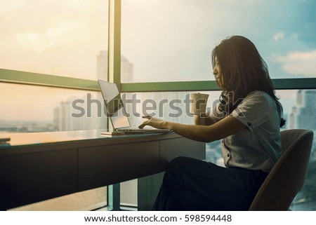 Relaxed Asian woman watching a laptop and holding a cup of coffee in background of river beautiful view at sunset and background blur building skyscrapers. (vintage color tone, advertise concept)