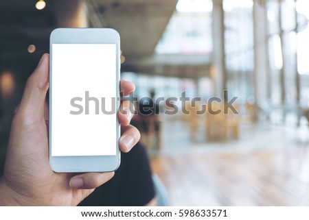 Mockup image of hand holding white mobile phone with blank white screen in modern cafe