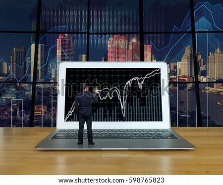 Businessman standing over the computer laptop on the wood table and writing the trading graph at the screen over the blurred photo of trading graph on cityscape background, Business trading concept