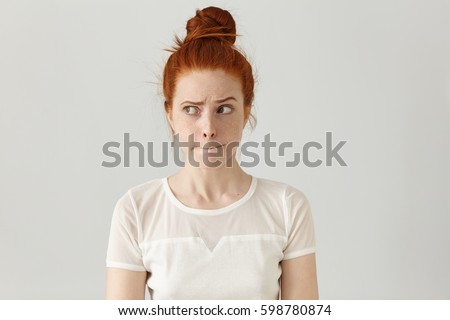 Indoor shot of cute redhead girl looking away, having doubtful and indecisive face expression, pursuing her lips as if forbidden to say anything. Confused young female posing isolated at white wall Royalty-Free Stock Photo #598780874