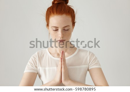 Gorgeous young Caucasian female wearing her ginger hair in bun, holding hands in namaste or prayer, keeping eyes closed while practising yoga and meditating at home alone, having calm look on her face