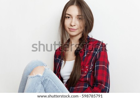 Indoor shot of attractive brunette woman teenager wearing her long hair in messy braid having carefree look wearing red checkered shirt over white t-shirt and ragged blue jeans, relaxing at home