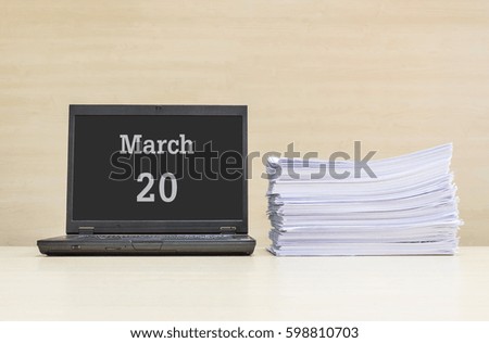 Closeup computer laptop with march 20 word on the center of screen in calendar concept and pile of work paper on wood desk and wood wall in work room textured background with copy space