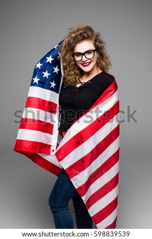 Cheerful young woman in casual clothes is holding the American flag and smiling while standing, on gray background