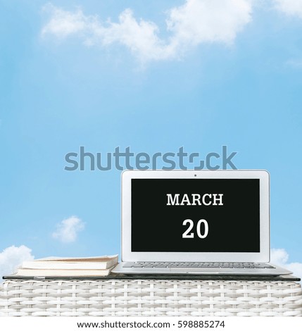 Closeup computer laptop with march 20 word on the center of screen in calendar concept on blurred wood weave table and book on blue sky with cloud textured background with copy space
