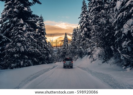 the car is riding on the powder snow road in the mountains with high trees on the gold sunset