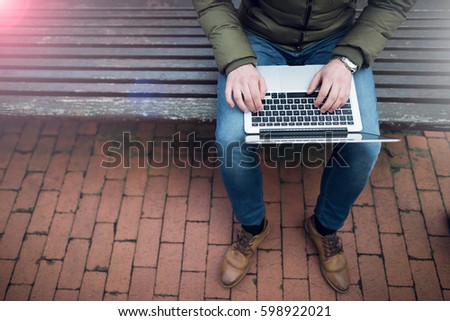 Laptop in a man's hands sitting on a wooden chair in park. Dressed in blue jeans and tan shoes. Photo with lens flare filter effect.