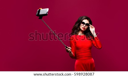 Portrait of beautiful brunette in awesome dress and red lips wearing sunglasses while making selfie with stick. Isolated