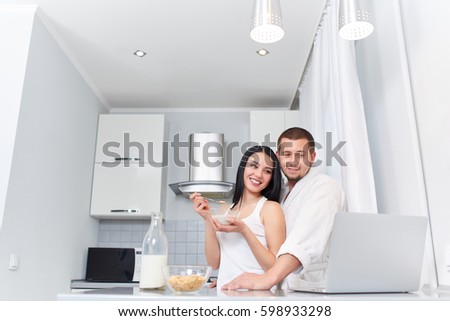 Morning of couple having breakfast at kitchen in morning, embracing and smiling together. Husband and woman eating oat meal with milk and watching funny video at laptop. Stylish kitchen interior.