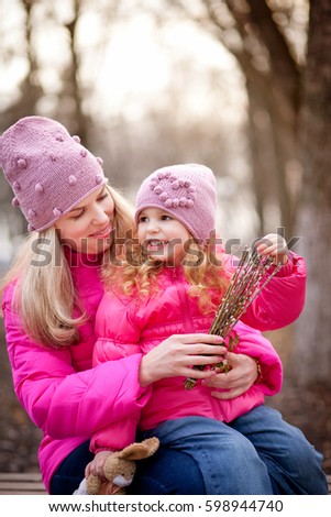 Blonde mom and little daughter in pink hats and jackets smile, hug and laugh outdoors in the spring for a walk in the park