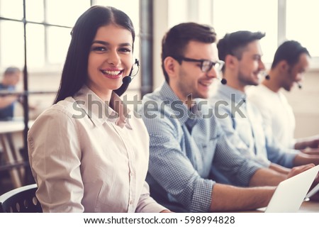 Beautiful business people in headsets are using computers and smiling while working in office. Girl is looking at camera Royalty-Free Stock Photo #598994828