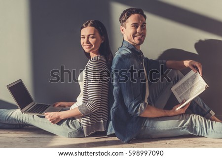 Beautiful young couple is looking at camera and smiling while sitting on the floor back to back. Girl is using laptop, guy is reading book