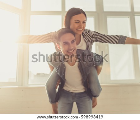 Beautiful young couple is smiling while standing near the window at home, girl is pickaback