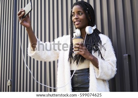 Attractive afro american young female blogger dressed in trendy casual outfit and with white headphones making selfie via smartphone to share with followers ideas for stylish look for city tour