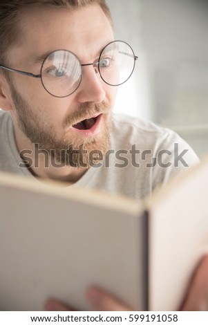 Closed up of surprised bearded young man wearing glasses sitting in home while reading books. Looking at book. Mouth opened.