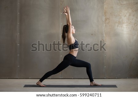 Young attractive woman practicing yoga, standing in Warrior one exercise, Virabhadrasana I pose, working out, wearing black sportswear, cool urban style, full length, grey studio background, side view Royalty-Free Stock Photo #599240711