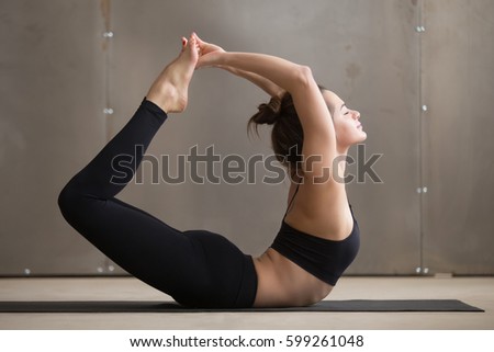 Young beautiful yogi woman practicing yoga, stretching in Dhanurasana exercise, Bow pose, working out, wearing black sportswear, cool urban style, full length, grey studio background, profile view 