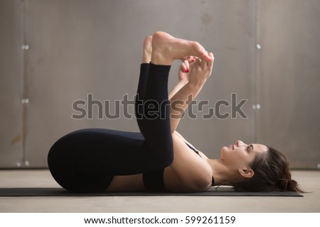 Young attractive woman practicing yoga, stretching in Ananda Balasana exercise, Happy Baby pose, working out, wearing black sportswear, cool urban style, full length, grey studio background, side view
