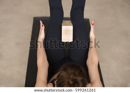 Young attractive woman practicing yoga using wooden block, doing navasana exercise, working out, wearing black sportswear, cool urban style, grey studio floor background, closeup, high angle view 