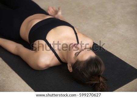 Young beautiful woman practicing yoga, lying in Dead Body, Savasana exercise, Corpse pose, working out, wearing black sportswear, cool urban style, closeup, grey studio background  Royalty-Free Stock Photo #599270627