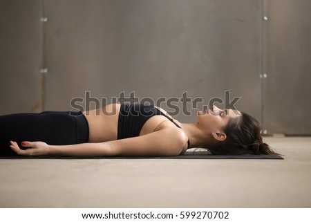 Young attractive woman practicing yoga, lying in Dead Body, Savasana exercise, Corpse pose, working out, wearing black sportswear, cool urban style, closeup, grey studio background  Royalty-Free Stock Photo #599270702
