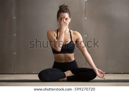 Young attractive sporty yogi woman practicing yoga, sitting in Sukhasana pose, Alternate Nostril Breathing, nadi shodhana pranayama exercise, working out, cool urban style, full length, grey studio  Royalty-Free Stock Photo #599270720