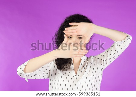 young curly brunette woman using her hands to create a border around her face on a pink background.