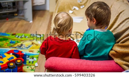 children room with two small kids brother and sister sit on burgundy sofa and reading book with pictures back view. Older brother show to small blonde sister pictures on soft book in homely atmosphere