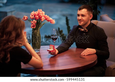 Young man makes woman marriage proposal and offers her jewelry case with engagement ring. They are sitting at table next to glasses of wine and vase with flowers. This is unexpected for her.