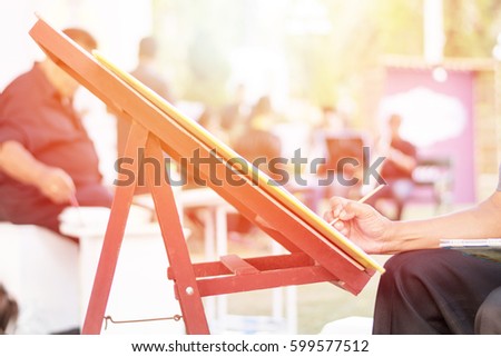 Hand of male artist painting picture on canvas in park with saxophone band playing in outdoor park.Jazz festival with saxophonist group on background for painter doing art work in public park holiday 