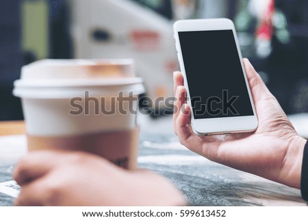 Mockup image of hands holding white mobile phone with blank black screen and hot coffee cup in modern cafe