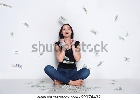 Excited woman sitting under money rain sit on floor and smile. Happy girl drop american dollar banknotes in the air - lottery jackpot and financial success concept, white studio background