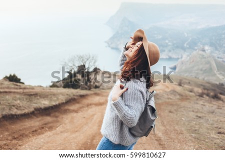 Girl wearing hat and sweater travel in mountains alone. Cold weather, calm scene. Backpacker walking outdoors in fall, back view over landscape. Wanderlust photo series.