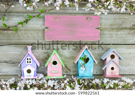 Blank pink wood sign hanging over colorful display of birdhouses and spring tree blossoms on rustic antique wooden background; springtime background with white flowers and copy space