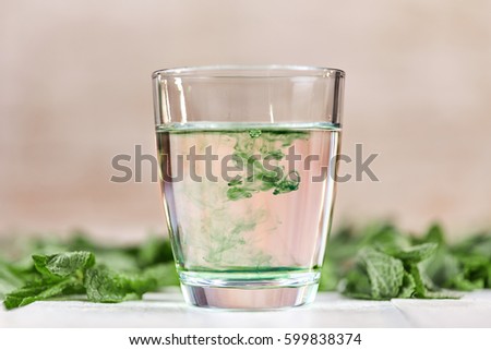 Dissolving liquid chlorophyll in glass of water on white table with green mint