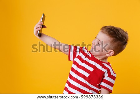 Portrait of a smiling little boy holding mobile phone and making selfie isolated over orange background