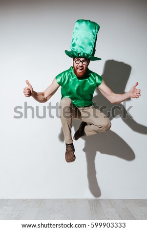 Full length of jumping happy man in green st.patriks costume which showing thumbs up, screaming and looking at camera. Isolated gray background Royalty-Free Stock Photo #599903333
