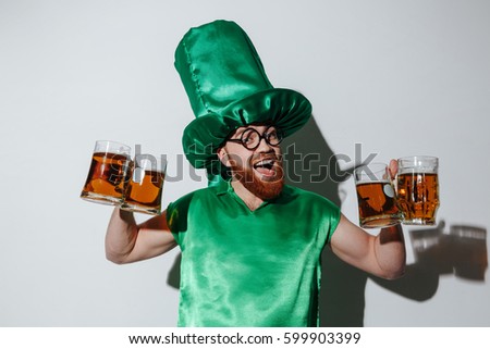 Bearded guy in st.patriks costume and eyeglasses which holding many cups and looking at camera. Isolated gray background