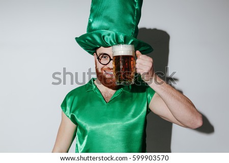 Pleased man in st.patriks costume and eyeglasses which holding cup of beer near the face and looking at camera. Isolated gray background