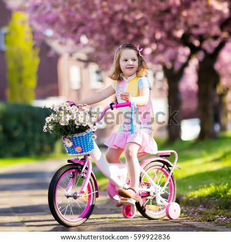 Child riding a bike on street with blooming cherry trees in the suburbs. Kid biking outdoors in urban park. Little girl on pink bicycle. Healthy preschool children summer activity. Kids play outside.