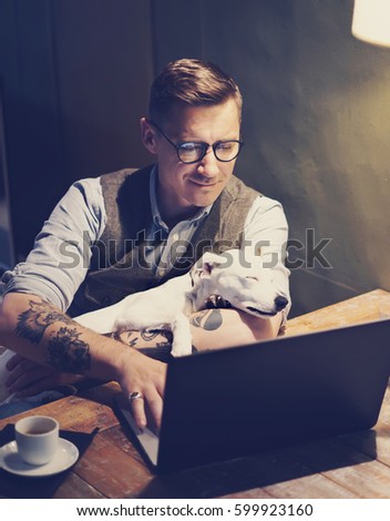 Smiling tattooed man in eyeglasses working at home on laptop while sitting at the wooden table with cute dog sleeping on his hands.Adult guy using modern computer for surfing web.Vertical