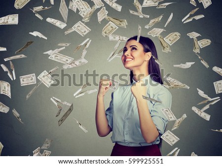 Portrait happy woman exults pumping fists ecstatic celebrates success under a money rain falling down dollar bills banknotes isolated on gray wall background with copy space  