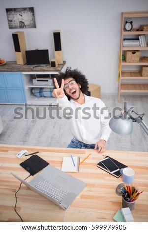 Hipster freelance man in glasses showing yo or okay sign to camera while sitting at table and working on laptop computer at home. Freelance or business concept.
