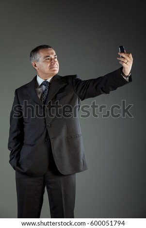 Studio shot of a senior man wearing elegant business suit while taking a selfie with the mobile phone against grey background