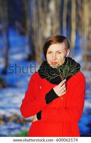Wonderful portrait of a cute young girl in a red coat with a green twig in the hands at sunset on blurred background of a winter forest