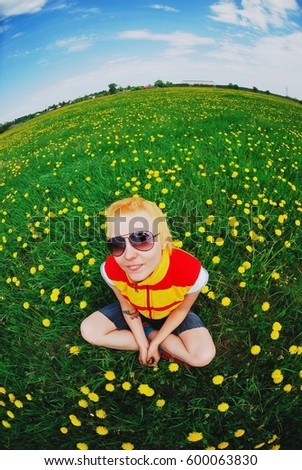 Portrait of young pretty smiling blonde with short hair in a yellow and red waistcoat in sunglasses, sitting in a clearing among yellow flowers of a dandelion. Beautiful skyline with fish-eye effect.