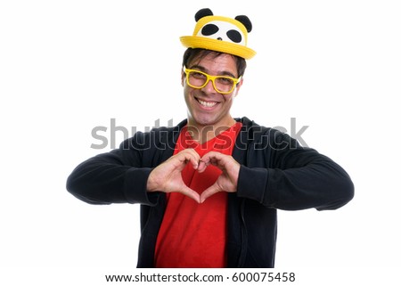 Studio shot of young happy Persian man smiling and giving hand heart sign while wearing cute hat and yellow eyeglasses isolated against white background