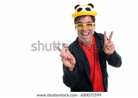 Studio shot of young happy Persian man smiling and giving peace sign with both hands while wearing cute hat and yellow eyeglasses isolated against white background