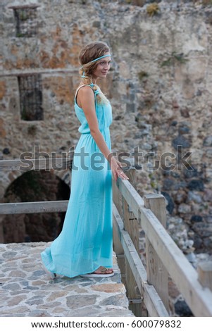 Young blonde woman in  turqoise maxi dress with braided hair against blurred stone castle wall