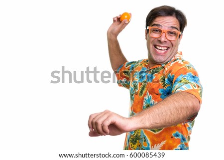 Studio shot of young happy Persian tourist man smiling and ready to throw orange fruit while wearing orange colored eyeglasses isolated against white background